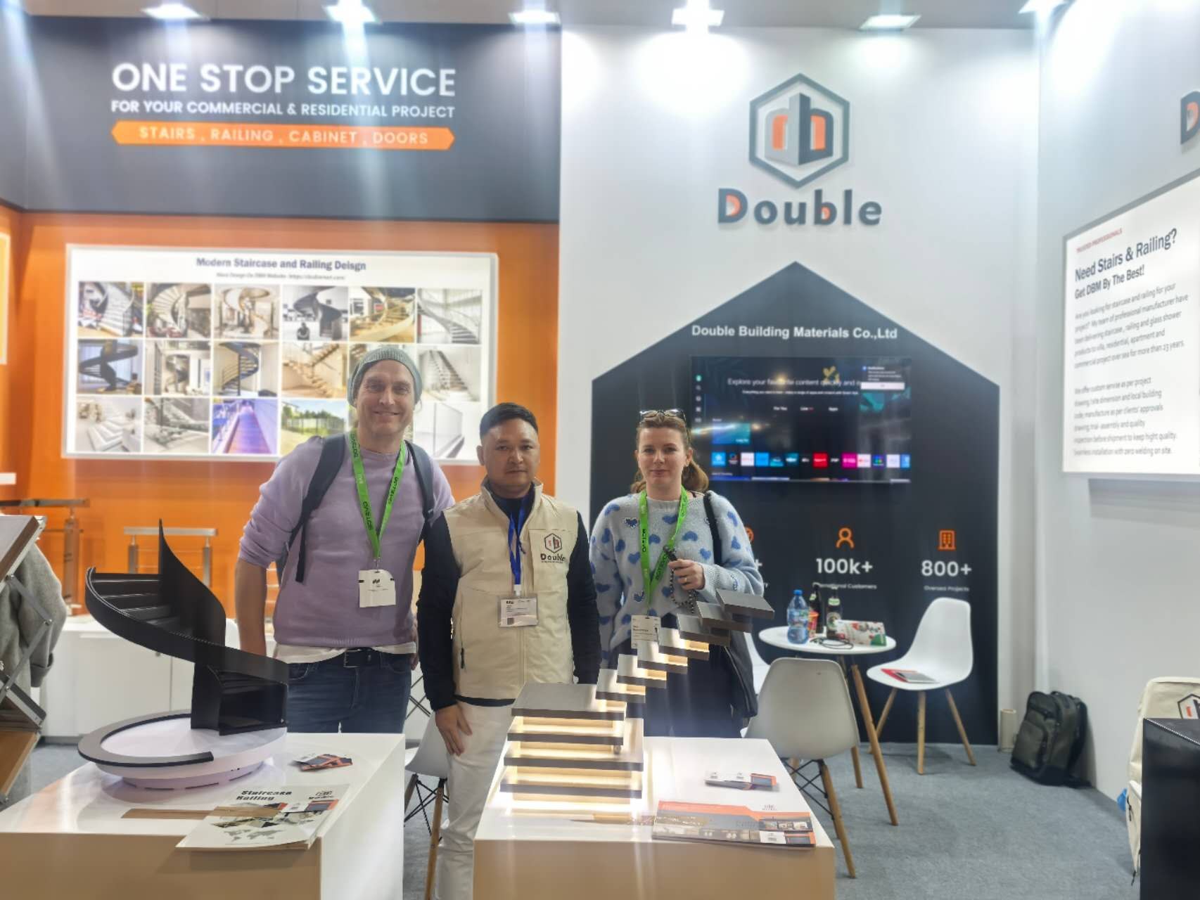 The Double Building Materials stand at BAU 2025 in Munich, with a one-stop-service wall, a spiral staircase model and a lit step sample, and team members with two visitors