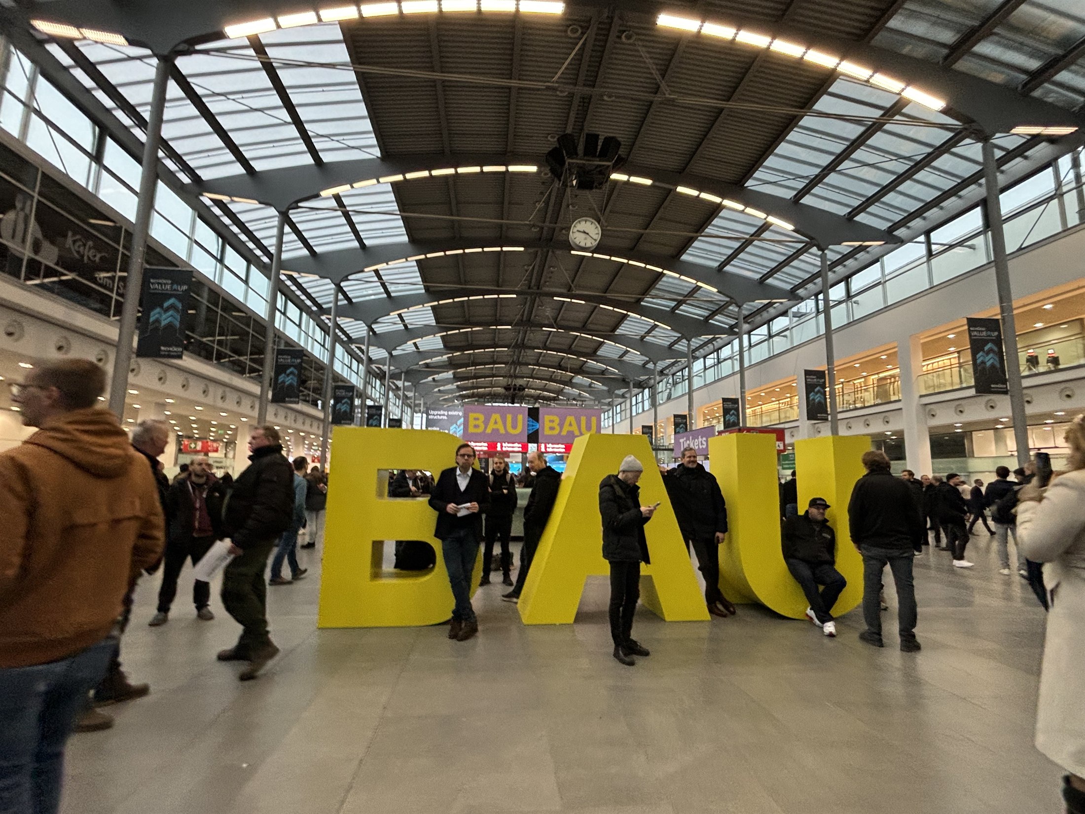 The large yellow BAU letters at the entrance of the Munich exhibition centre, with crowds of trade visitors walking past under the glass roof