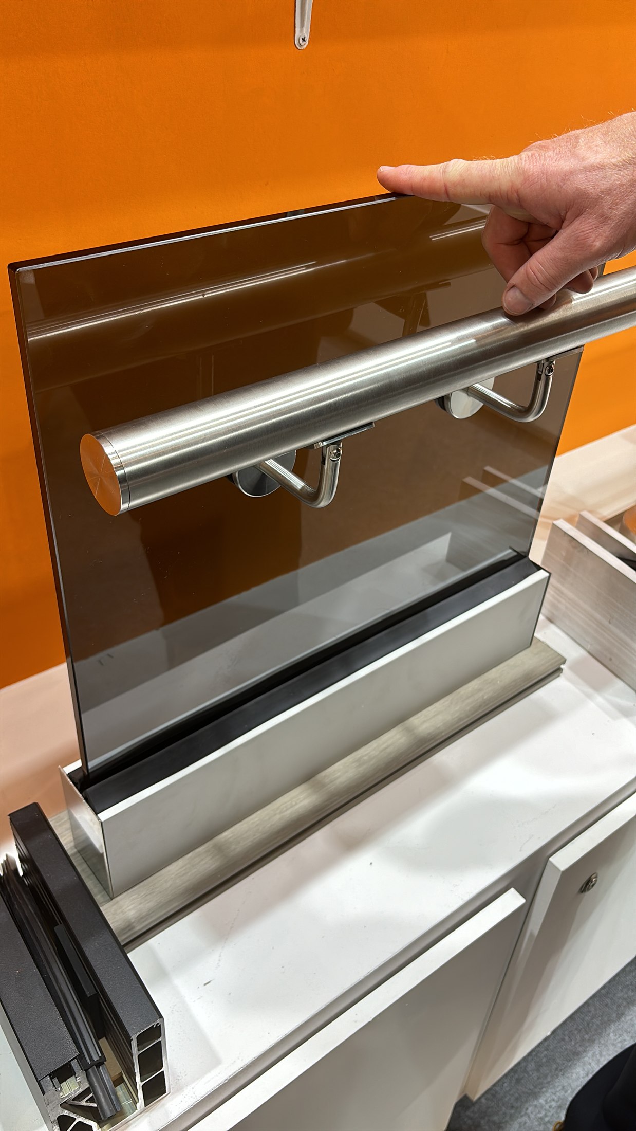 A close detail of a glass railing panel with a round stainless steel handrail and an aluminium base channel, shown as a sample at BAU 2025