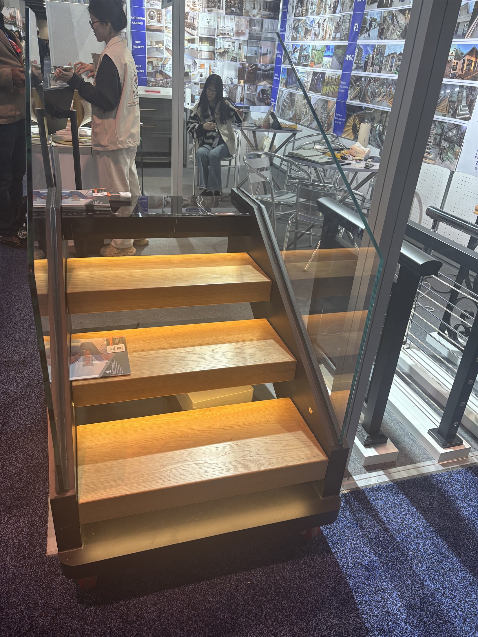 A floating wood-tread staircase model with a frameless glass balustrade and under-tread lighting shown at the IBS 2025 Las Vegas booth