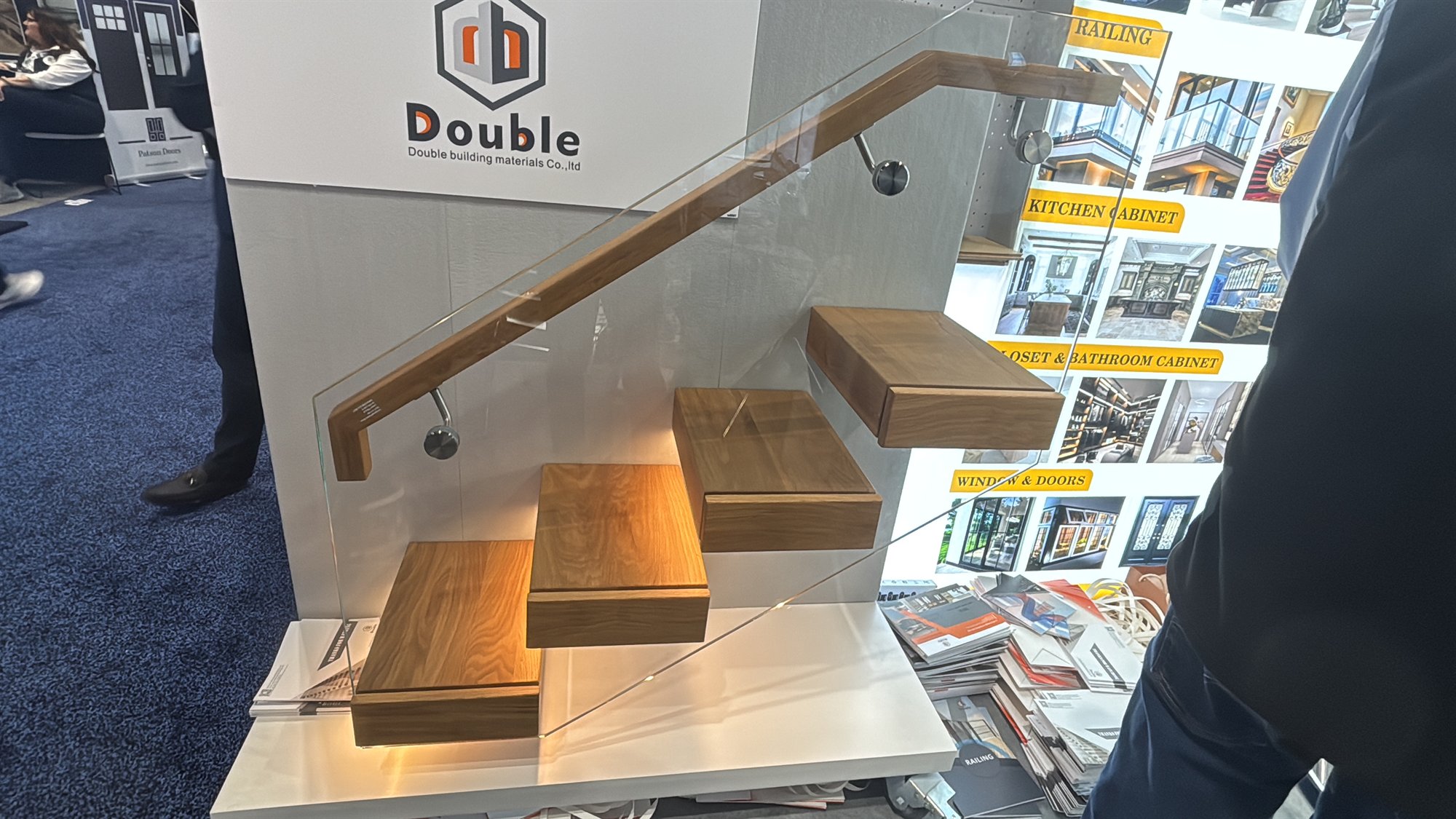 Floating staircase model with cantilevered oak treads, a frameless glass balustrade and a wood handrail, lit from under the treads at the Double Building Materials IBS 2026 booth