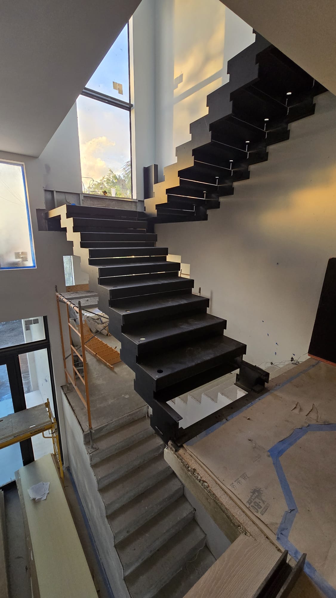 Matte-black steel stringer and welded tread plates of the floating switchback staircase, installed before the white oak goes on, Florida