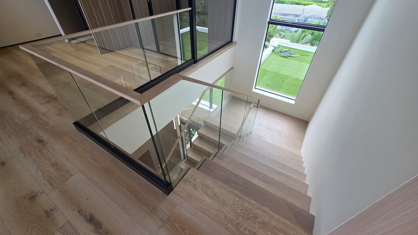 Top-down view of the finished white-oak floating staircase and half-landing, frameless glass with an oak cap, over a lawn and ocean view, Florida