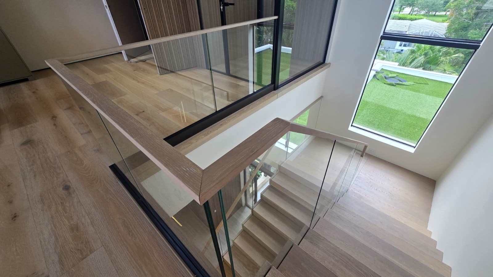 Overhead view of the white-oak half-landing and oak-capped glass balustrade, looking out to a lawn and palms in Florida