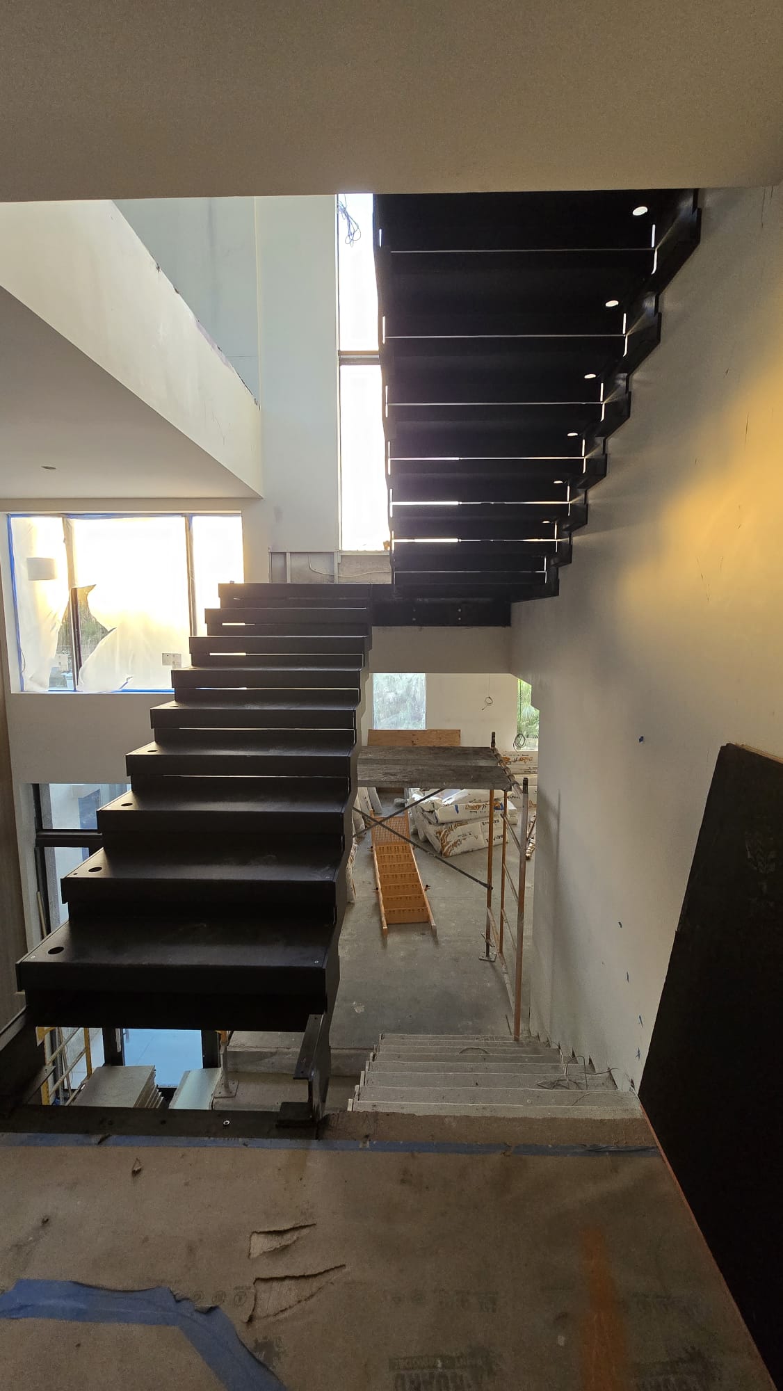 Underside view of the black-steel floating staircase frame during installation, showing the cantilevered tread plates and recessed soffit lights, Florida
