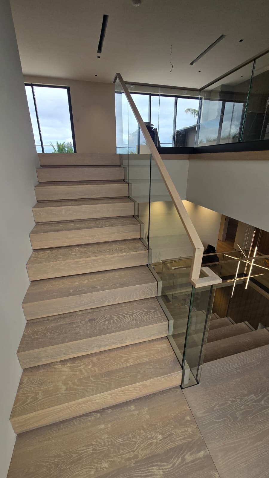 White-oak floating staircase with a glass balustrade and oak handrail, with the ocean visible through tall upper windows in a Florida home