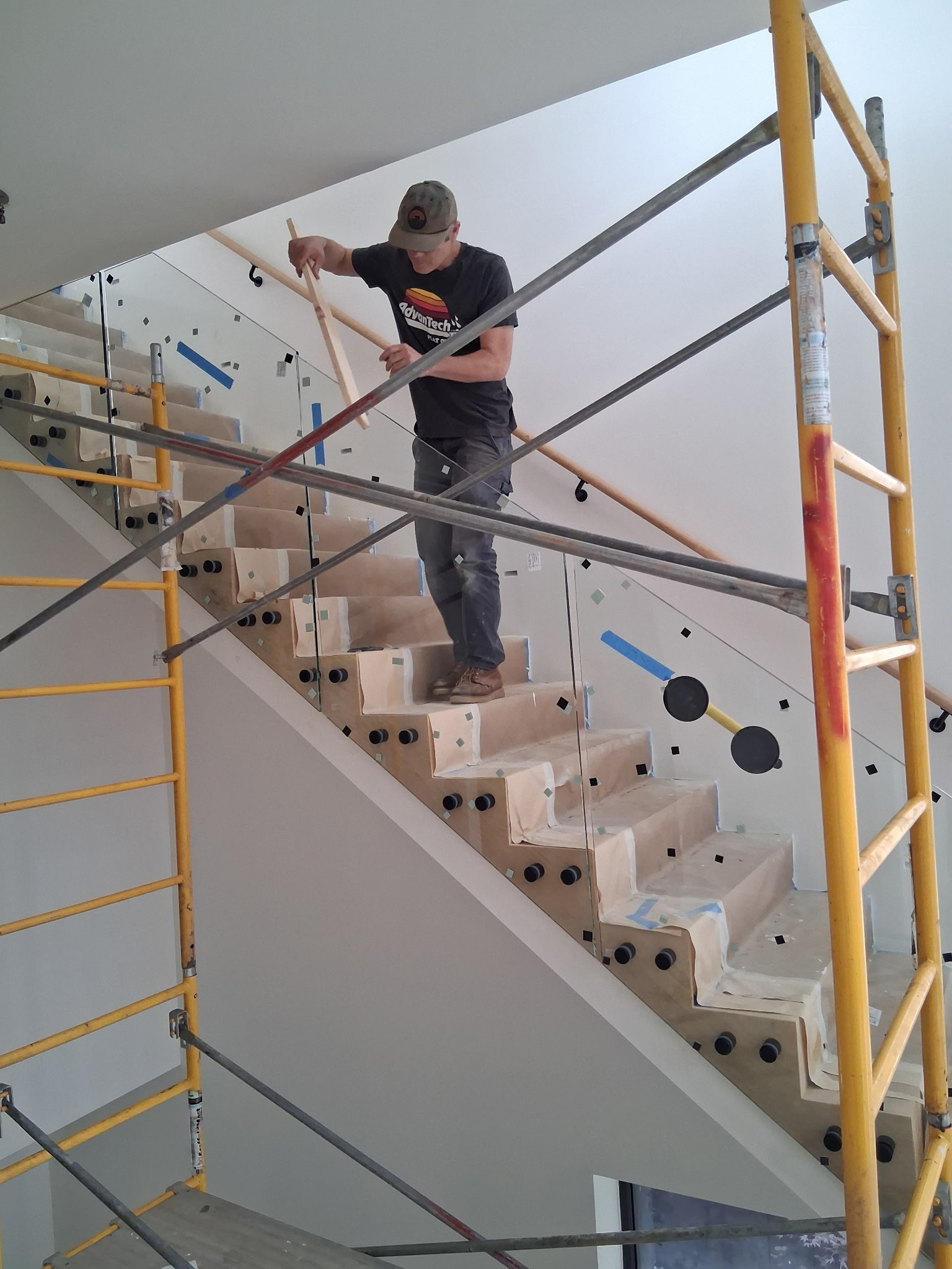 Standoff glass railing being set on an Atlanta staircase from a scaffold, two black pins at each paper-wrapped tread end