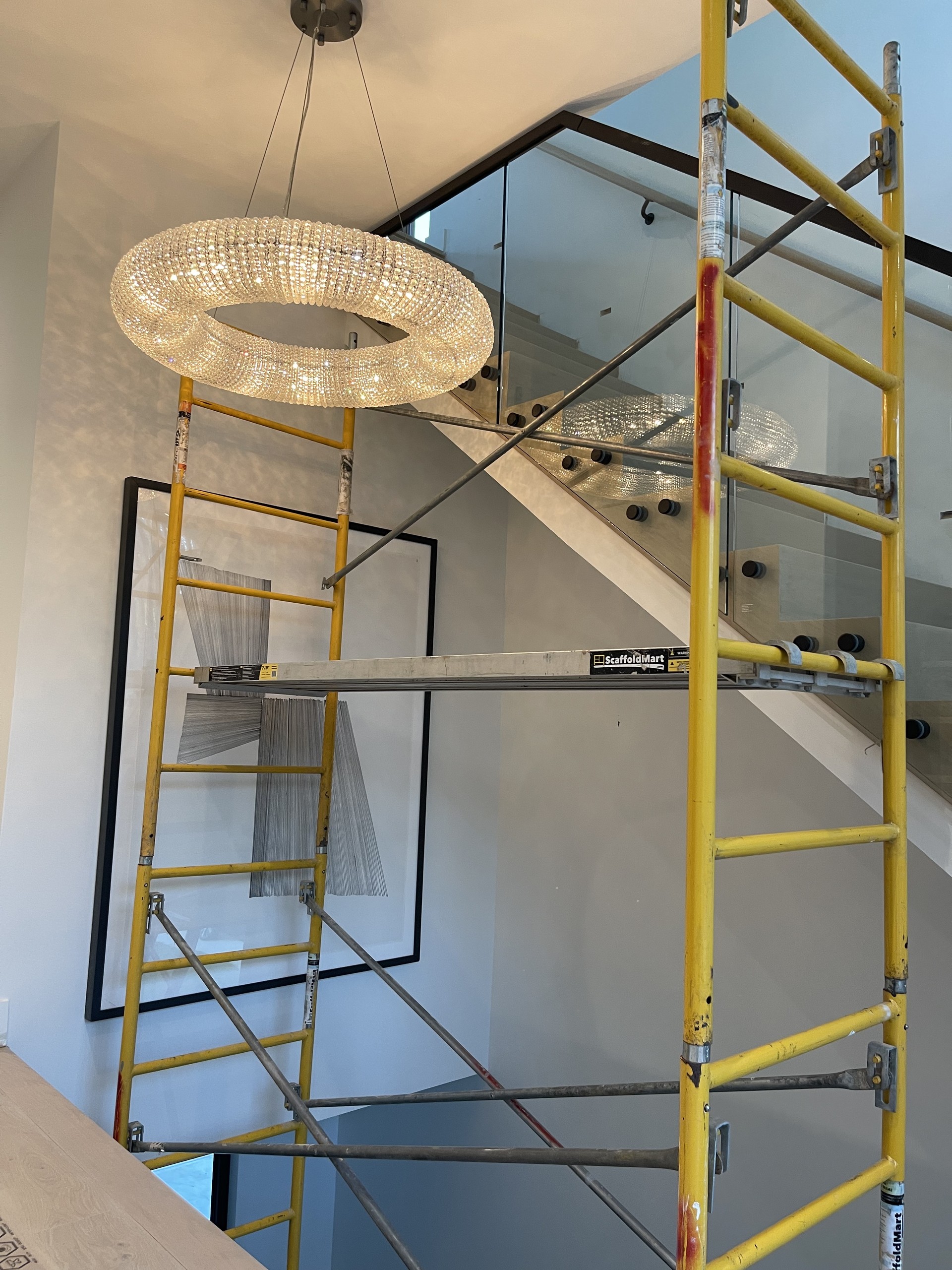 Glass stair railing with black standoff pins and a black top rail below a crystal ring chandelier, scaffold still in place