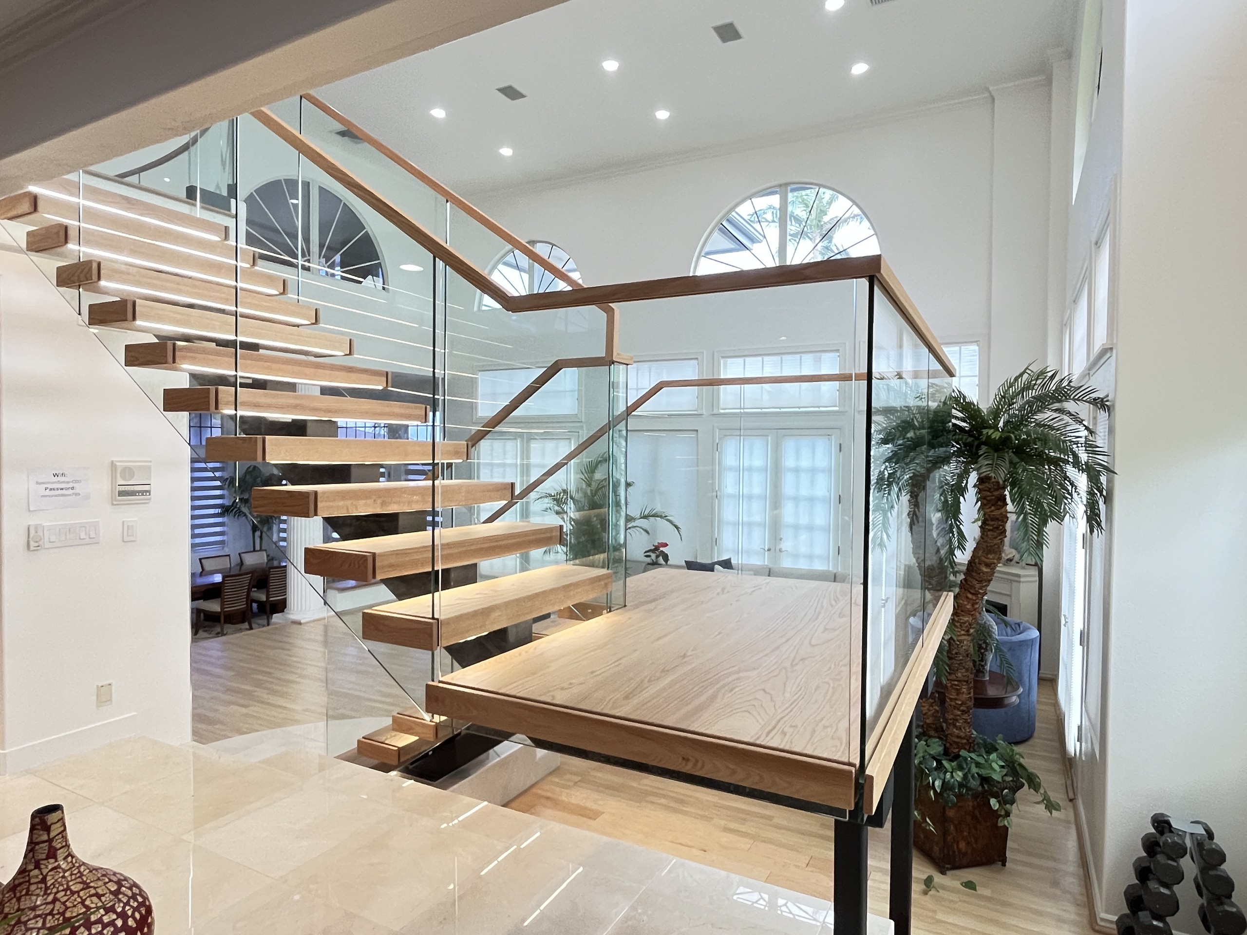 Red oak U-shaped staircase landing on black steel posts, wrapped in frameless glass with a wood handrail, in a Hawaii home with arched windows